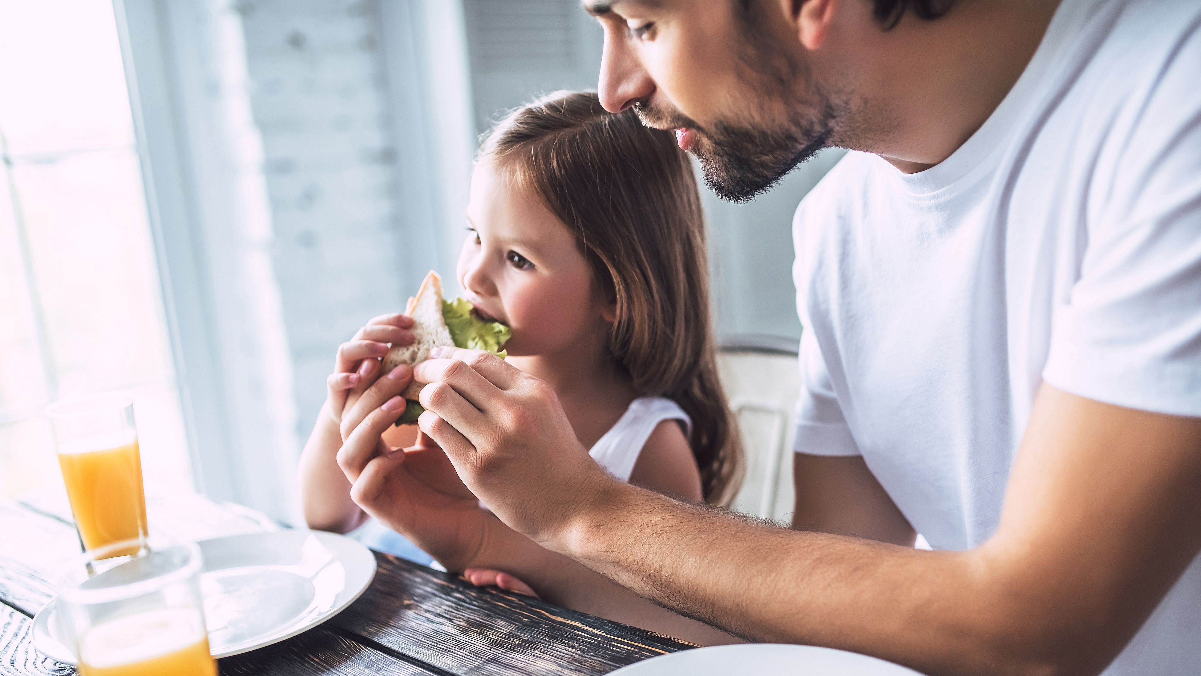 Die Aufnahme zeigt einen Vater mit seiner kleinen Tochter beim Frühstück. Der Vater hilft der Tochter beim Essen, indem er ihr das Sandwich hält.